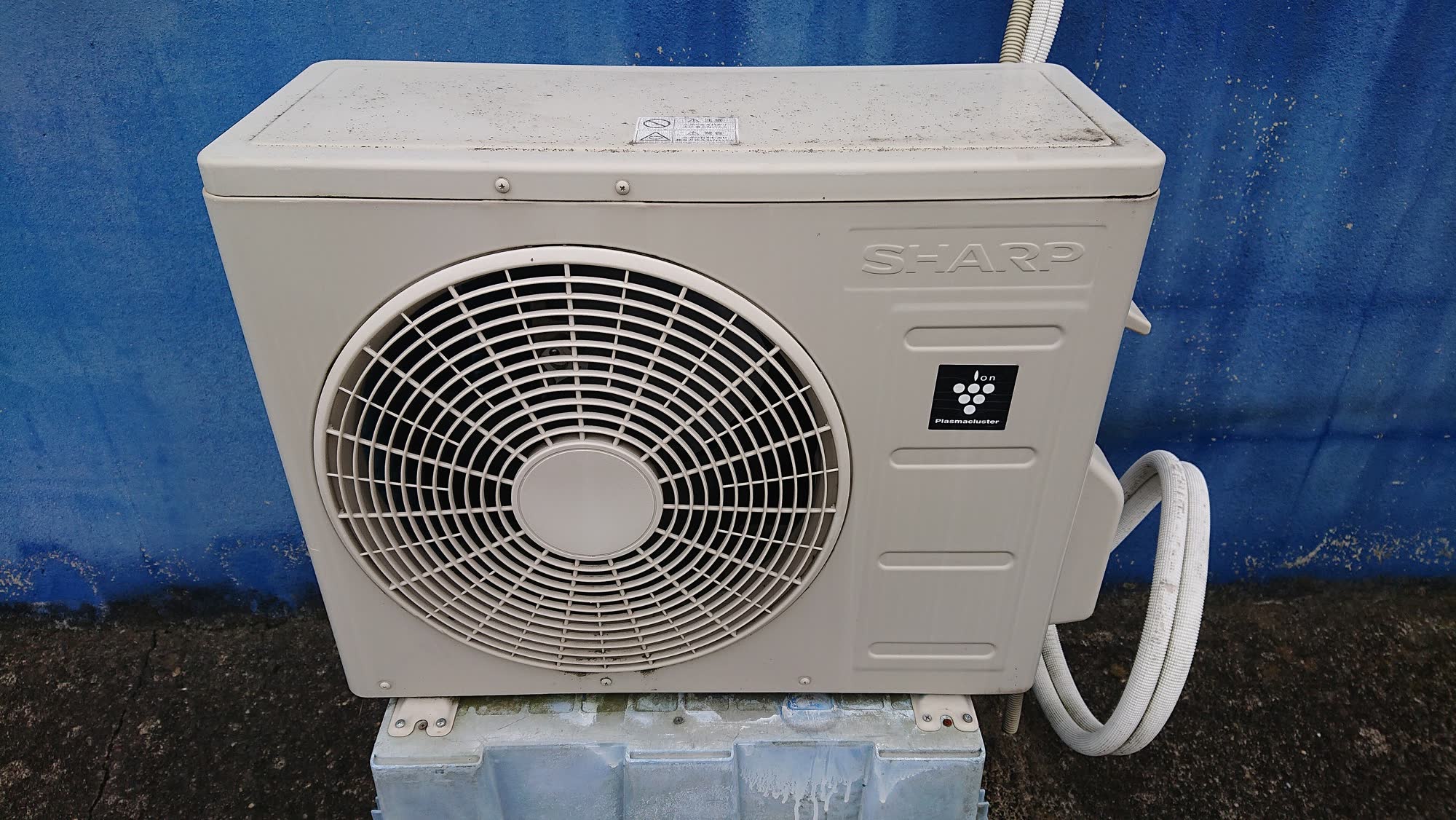 Outdoor air conditioner condenser unit with a large fan grille against a blue wall
